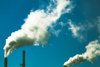 The image shows two industrial chimneys polluting the blue sky with thick, white plumes of smoke.
