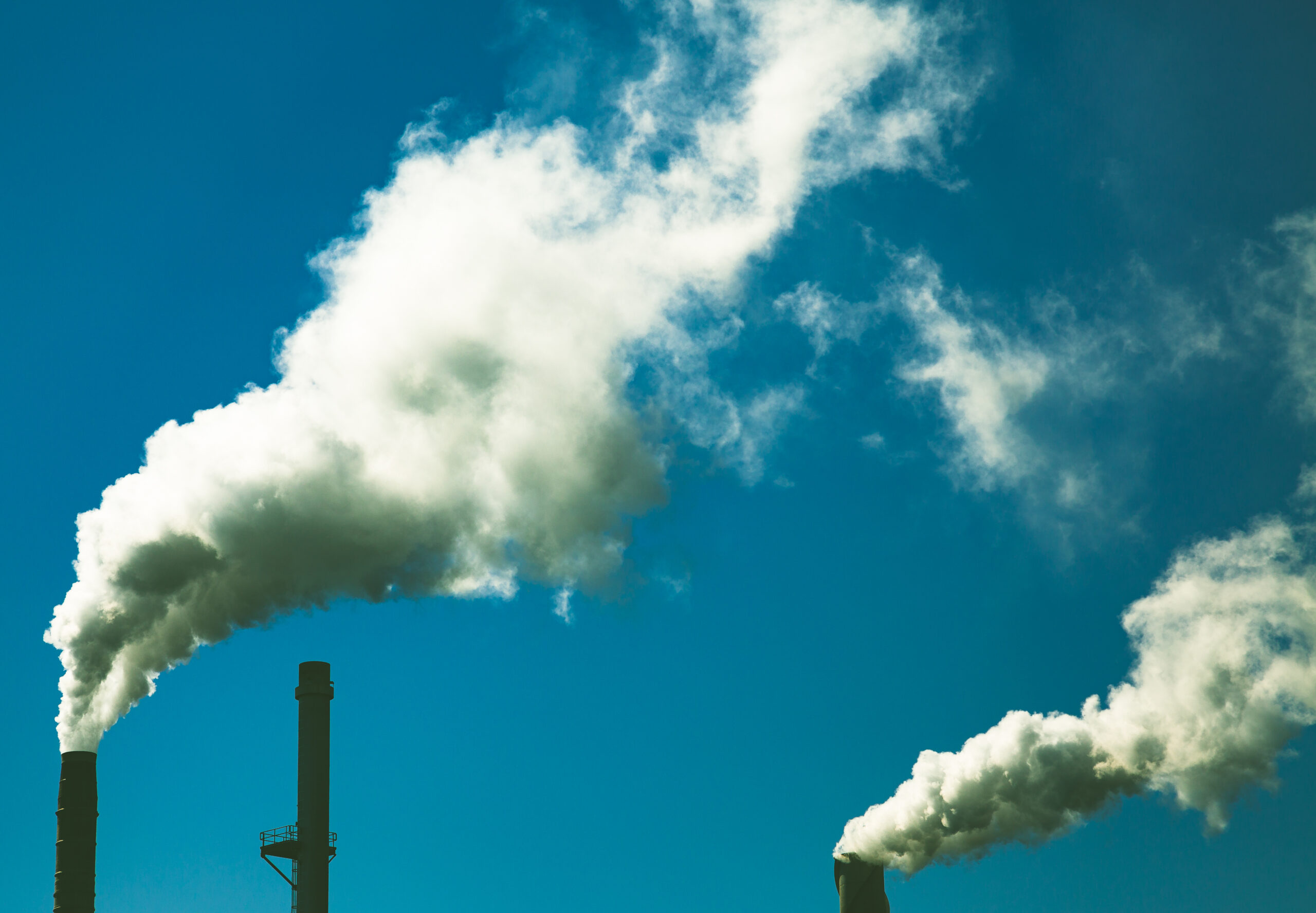 The image shows two industrial chimneys polluting the blue sky with thick, white plumes of smoke.