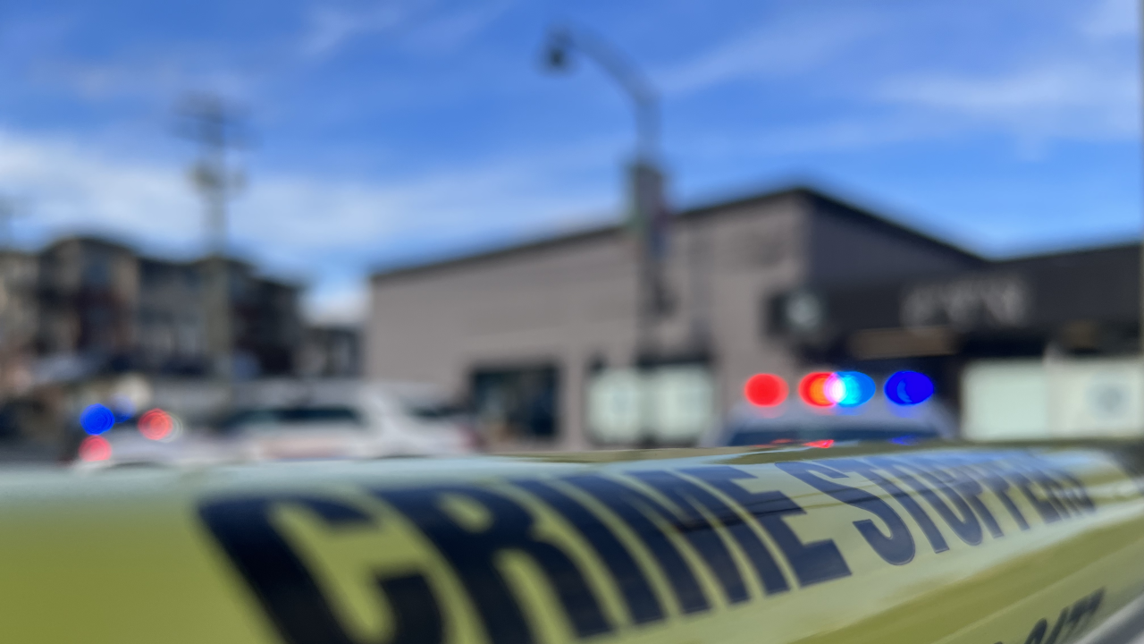 Yellow police tape in the foreground with blurred emergency vehicles and flashing lights in the background at a crime scene in British Columbia.