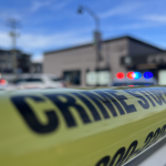 Yellow police tape in the foreground with blurred emergency vehicles and flashing lights in the background at a crime scene in British Columbia.
