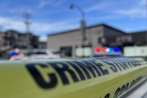 Yellow police tape in the foreground with blurred emergency vehicles and flashing lights in the background at a crime scene in British Columbia.