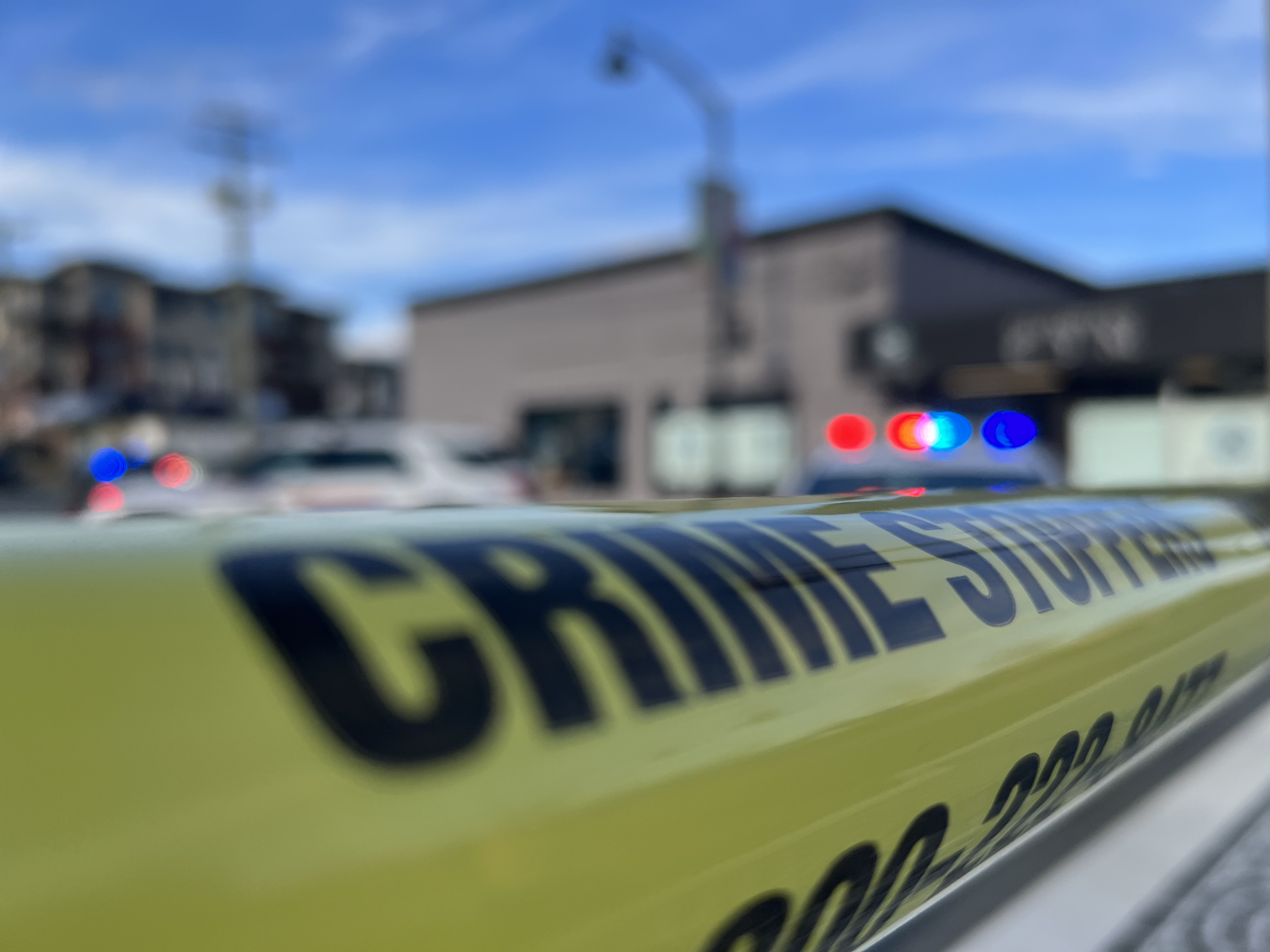 Yellow police tape in the foreground with blurred emergency vehicles and flashing lights in the background at a crime scene in British Columbia.
