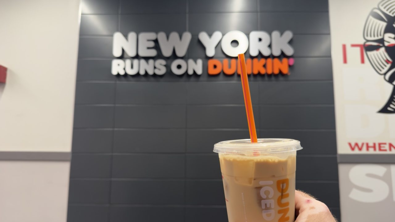 A Dunkin’ iced coffee held inside a New York City Dunkin’ location with a “New York Runs on Dunkin’” sign in the background.