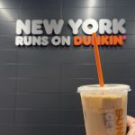 A Dunkin’ iced coffee held inside a New York City Dunkin’ location with a “New York Runs on Dunkin’” sign in the background.