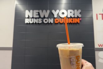 A Dunkin’ iced coffee held inside a New York City Dunkin’ location with a “New York Runs on Dunkin’” sign in the background.