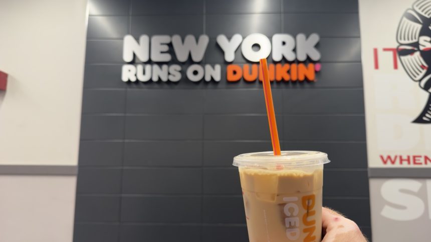 A Dunkin’ iced coffee held inside a New York City Dunkin’ location with a “New York Runs on Dunkin’” sign in the background.